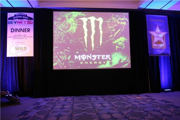 A projection screen on a dark ballroom stage shows the Monster Energy logo, with a Wild-sponsored BevNET Live dinner banner on the left and a Best of 2009 banner on the right.
