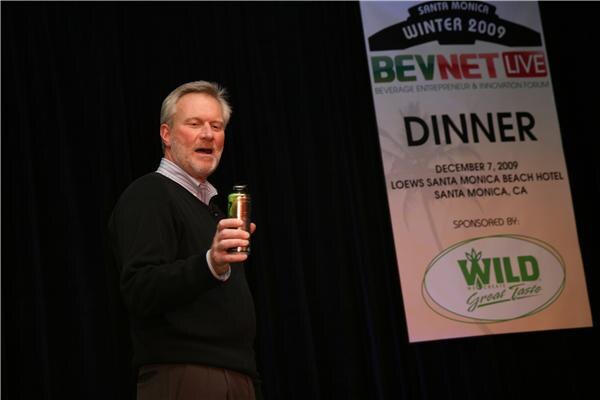 A man in a dark sweater holds up a canned beverage and speaks on stage beside a BevNET Live dinner banner sponsored by Wild, dated December 7, 2009.