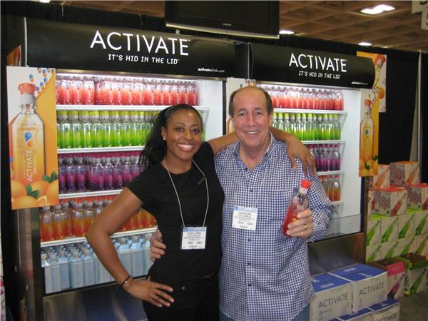 A smiling woman in a black top and a man in a blue checked shirt holding a red drink bottle pose with arms around each other in front of lit Activate drink coolers.