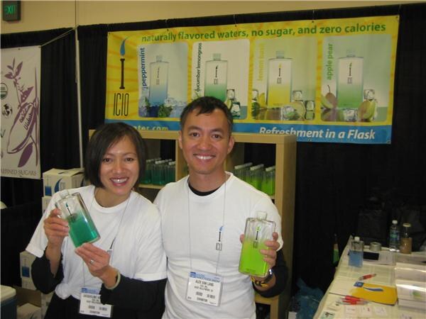 Alex Van Lang, center, and a smiling woman hold flask-shaped bottles of green Icio flavored water in front of an Icio banner listing peppermint, cucumber lemongrass, lemon basil and apple pear.