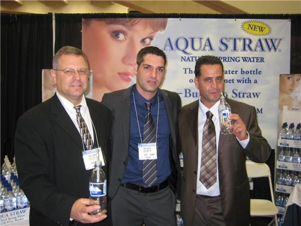 Three men in suits stand in front of an Aqua Straw natural spring water banner; two hold bottles of water and all wear show badges.