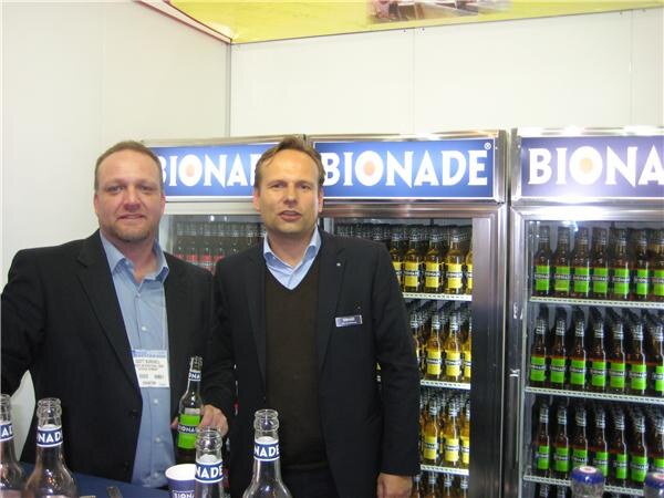 Two men in dark jackets stand behind a counter holding Bionade bottles, in front of glass coolers stocked with bottles under Bionade signs.