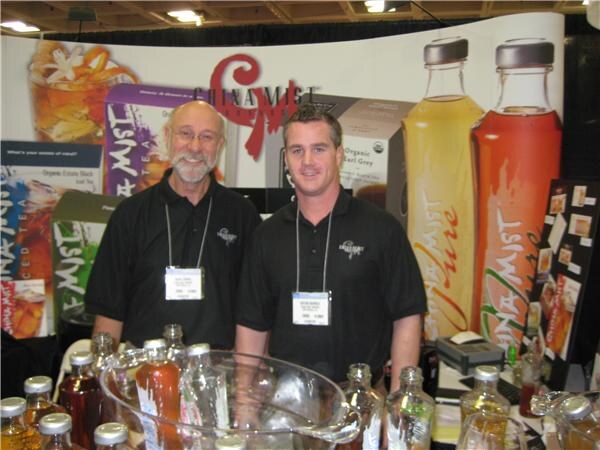 Two men in black China Mist polo shirts and show badges stand in front of tea packaging and bottled drink graphics, with bottles on the table.
