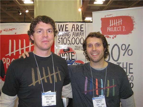 Darren Dunham of Project 7, Inc. on the left and Tyler Merrick of Project 7, Inc. on the right stand side by side wearing badges, in front of Project 7 banners.