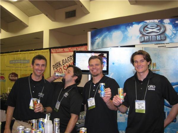 Four men in black Ex Drinks shirts hold energy drink cans at a booth with an Ex Drinks sign and a Pork Barrel BBQ banner behind them.