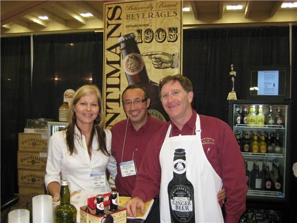 A woman and two men stand at a Fentimans booth; one man wears an apron printed with a Ginger Beer bottle. Fentimans banners, boxes and a cooler are behind them.