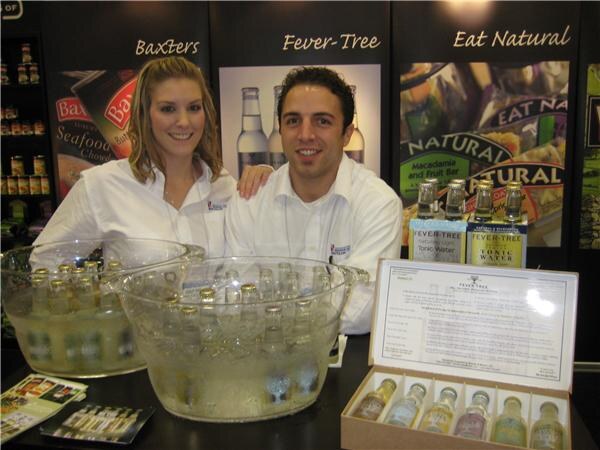 A smiling woman and man in white shirts stand behind two glass bowls of iced bottles at a booth with Baxters, Fever-Tree and Eat Natural signs, with Fever-Tree tonic water bottles beside them.