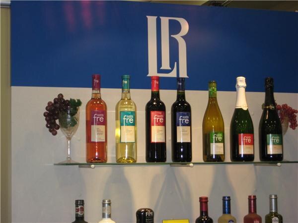 Seven bottles of Fre wine stand on a glass shelf below a blue and white wall with an LR logo, with a decorative grape cluster and a wine glass at the left end.