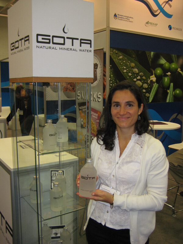 A smiling woman with dark wavy hair and a white cardigan holds a Gota bottle beside a glass display case of Gota natural mineral water bottles under a large Gota sign.