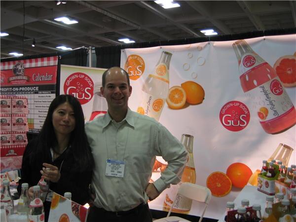 A woman with long dark hair and a smiling man in a light green shirt with a badge stand in front of a Grown-Up Soda GuS banner showing oranges and bottles, with soda bottles on the table.