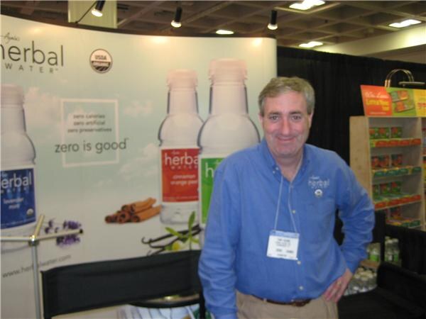 A man in a blue shirt with a show badge stands in front of a Herbal Water backdrop showing bottles and the words "zero is good".
