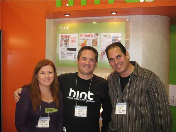 A woman and two men with badges pose together at a Hint booth with an orange wall; one man wears a black Hint shirt reading Drink water, not sugar, and magazine features are displayed behind.