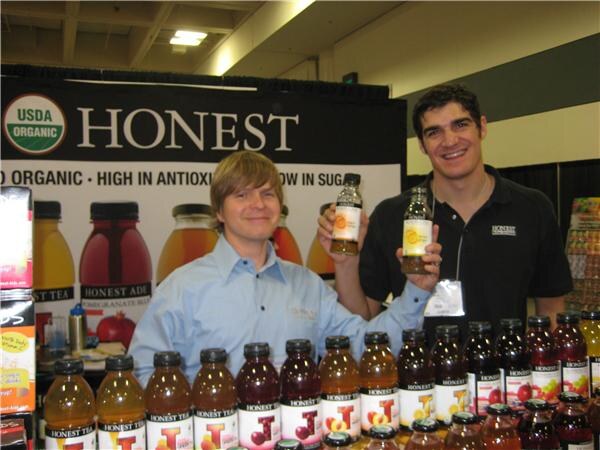 Two smiling men hold up bottles of Honest Tea behind rows of Honest Tea and Honest Ade bottles, in front of a USDA Organic Honest banner.