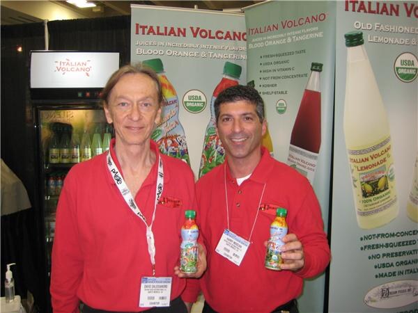 Two men in red shirts hold small bottles of juice in front of Italian Volcano banners advertising blood orange, tangerine and lemonade drinks.