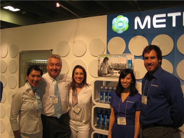 Five smiling people with badges, three women and two men, stand in front of a Metromint booth with a Metromint Electro display of bottled water on white shelves.