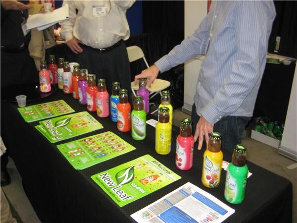 A table with black cloth displays a row of colorful New Leaf tea bottles and green New Leaf flyers. A man in a striped shirt reaches toward the bottles, and another person stands behind.