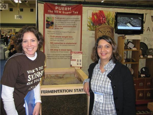 Two smiling women stand beside a Numi Tea fermentation station display with a Puerh the new super tea sign; one wears a brown shirt reading What's the skinny on puerh.