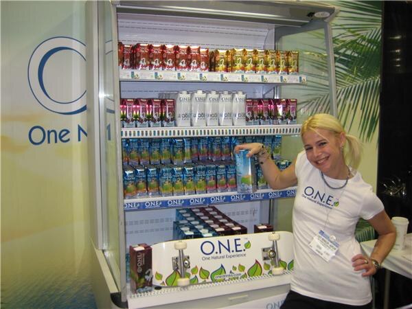 A smiling blonde woman in an O.N.E. shirt leans on an open refrigerated case stocked with O.N.E. drink cartons, with a skateboard deck on the lower shelf.