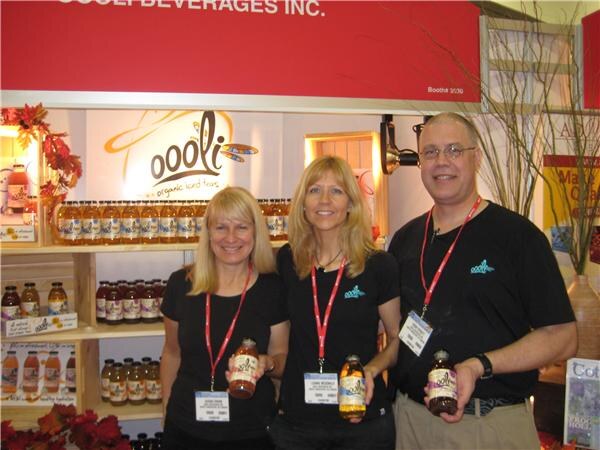 Two women and a man in black shirts and badge lanyards stand in the Oooli Beverages Inc. booth, each holding a bottle of iced tea, with shelves of bottles behind them.