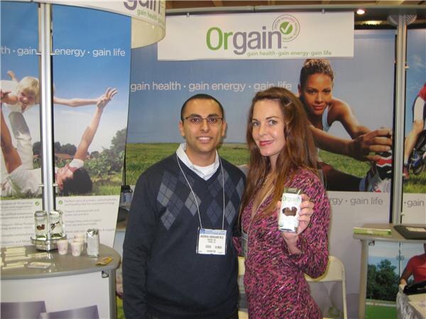 A man in a dark argyle sweater and a woman in a pink patterned top stand in the Orgain booth. She holds a small Orgain container. Banners behind them read gain health, gain energy, gain life.