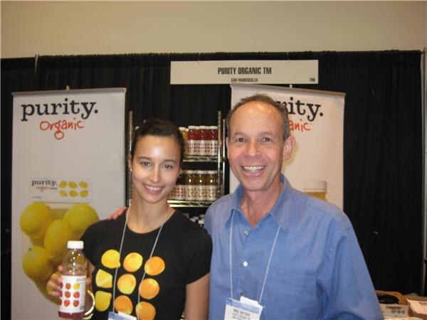 A smiling woman in a black lemon-print shirt holding a bottle and a smiling man in a blue shirt stand in the Purity Organic booth, with Purity Organic banners and bottles on shelves behind them.