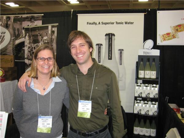 A woman with glasses and a man in a green shirt stand with arms around each other in the Q Tonic booth, in front of a banner reading Finally, A Superior Tonic Water and a shelf of tonic bottles.