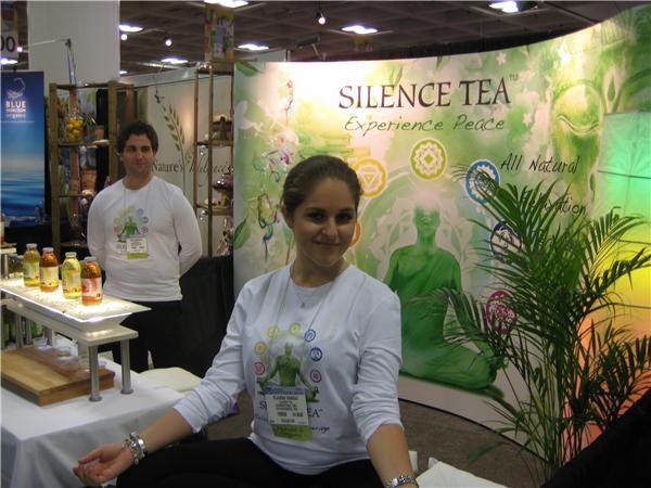 A woman in a white long-sleeved shirt sits in front of a green Silence Tea banner. A man in a white shirt stands behind her, and bottles of tea sit on a table at left.