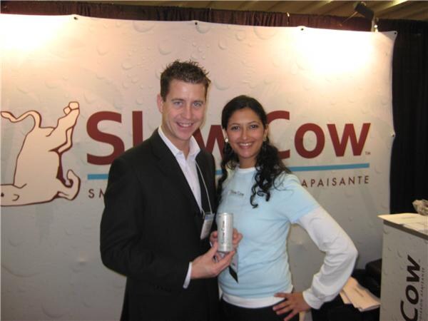 A man in a dark suit holding a silver can and a woman in a light blue top stand in front of a Slow Cow banner with a cow outline.