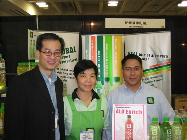 Three people stand in front of an SPI West Port, Inc. sign and ALŌ aloe vera banners, with green-labeled bottles and an ALŌ Enrich poster in front.