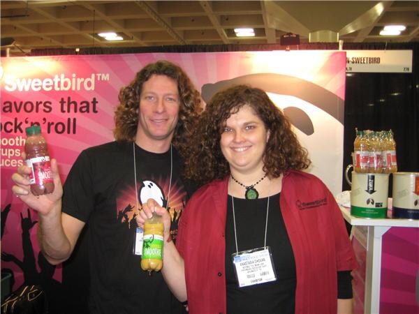 A curly-haired man in a black T-shirt and a woman in a red jacket hold bottles of Sweetbird smoothies in front of a pink Sweetbird banner.
