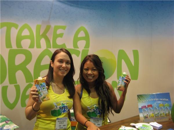 Two smiling women in yellow Vita Coco tank tops hold up Vita Coco cartons in front of a green banner reading "Take a hydration vacation"; a product poster sits on the table.