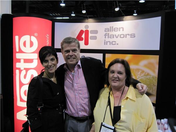 A man in a pink striped shirt stands between two women in front of an Allen Flavors Inc. sign and a red Nestlé banner, with his arm around the woman at right.