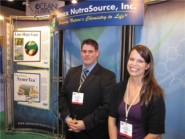 A man in a dark suit and a smiling woman in a purple top, both wearing badges, stand in the Amax NutraSource booth. Posters show SynerTea and Luo Han Guo with P&G logos.