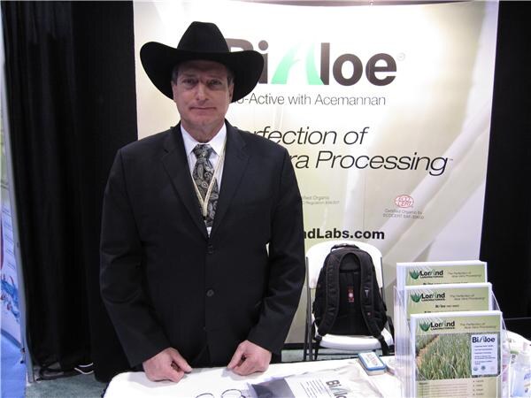 A man in a black cowboy hat, dark suit and patterned tie stands behind a table at the BiAloe booth, with brochures and a backpack beside him and a BiAloe banner behind him.
