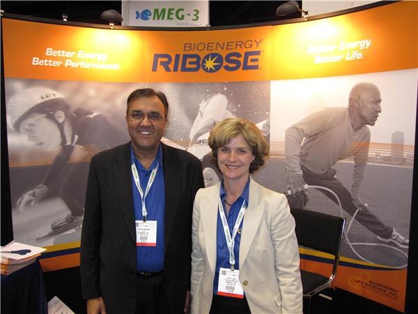 A man in a dark blazer and a woman in a cream jacket, both wearing lanyards, stand in front of orange BioEnergy Ribose banners showing athletes. A MEG-3 sign hangs above.