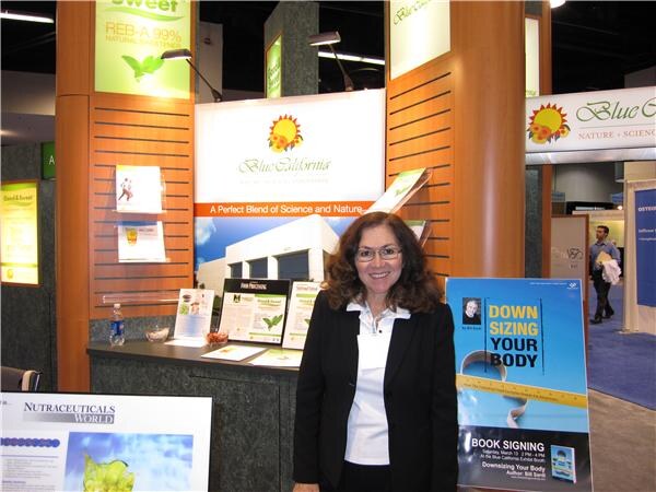 A woman with glasses and a dark blazer smiles in front of the Blue California booth. A Nutraceuticals World sign and a "Downsizing Your Body" book signing poster stand nearby.
