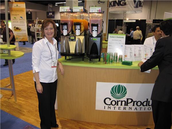 A woman in a white shirt and black pants stands beside a CornProducts International counter holding three beverage dispensers and printed handouts; a man in a dark suit stands at right.