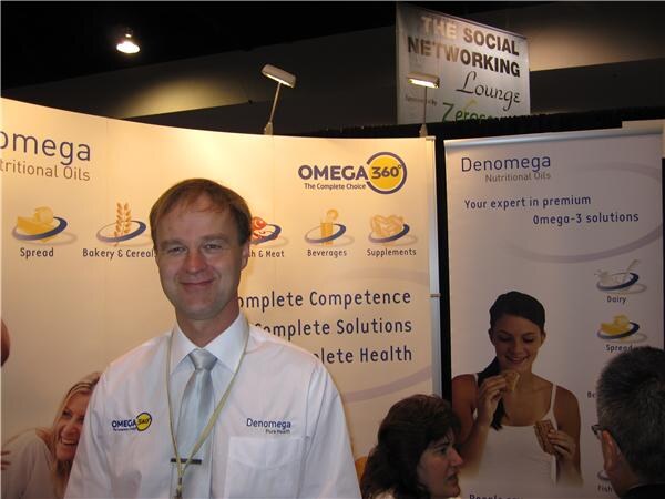 A smiling man in a white Denomega shirt and gray tie stands in front of Omega 360 and Denomega Nutritional Oils banners, with a Social Networking Lounge sign overhead.