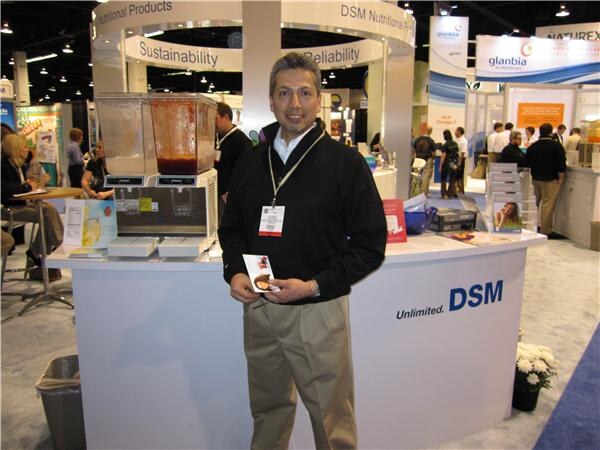 A man in a black jacket and tan pants with a lanyard badge holds a brochure in front of a curved DSM booth counter with a beverage dispenser, DSM Nutritional Products signs and Glanbia banners behind.