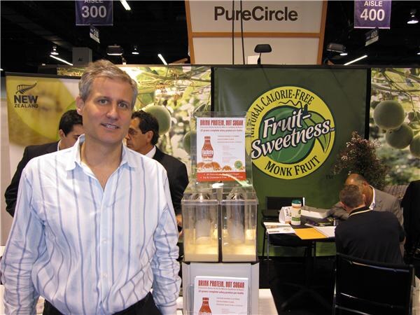 A man in a light blue striped shirt stands in front of a green Fruit-Sweetness monk fruit banner under a PureCircle sign, beside a display of protein drink bottles; other people sit and stand behind him.