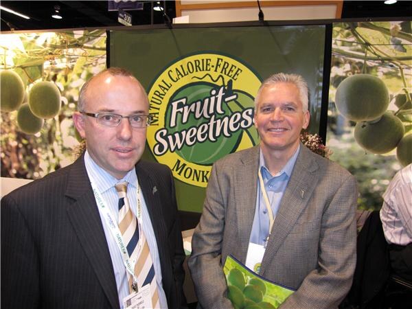 Two men in suit jackets stand side by side in front of a green Fruit-Sweetness natural calorie-free monk fruit banner; one wears glasses and a striped tie, and both wear show badges.
