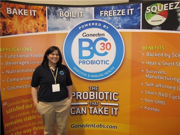 A smiling woman in a navy polo and khaki pants with a lanyard stands before an orange Ganeden BC30 Probiotic backdrop reading 'The probiotic that can take it', with Bake it, Boil it, Freeze it panels.