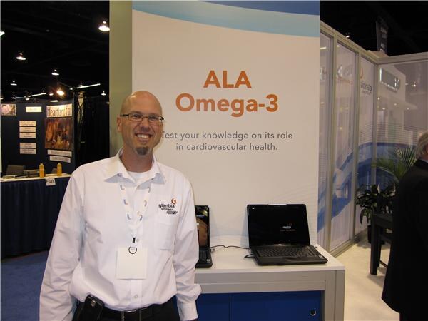 A smiling bald man with glasses and a goatee, wearing a white shirt with a Glanbia logo and a lanyard, stands beside a display reading 'ALA Omega-3' with two laptops on the counter.