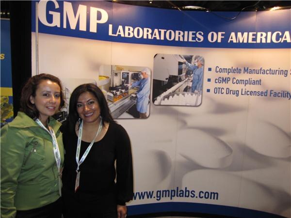 Two smiling women with lanyards stand in front of a blue and white GMP Laboratories of America backdrop listing manufacturing capabilities, with photos of workers in protective clothing.