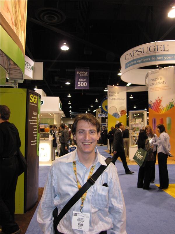 Gregory Drew of Pharmachem smiles in a checked shirt with a lanyard and shoulder strap on an aisle, with Capsugel and SGF booth signs and other attendees behind him.