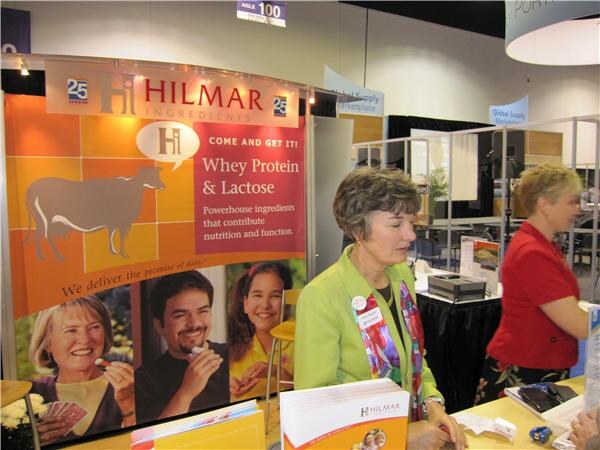 Two women stand at a counter in the Hilmar Ingredients booth, which has a banner reading 'Whey Protein & Lactose'. One wears a green jacket and badge, the other a red top. Brochures lie on the counter.
