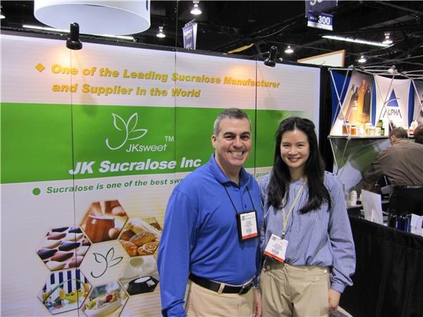 A smiling man in a blue polo and a smiling woman in a striped shirt, both with badges, stand in front of a green JK Sucralose Inc JKsweet banner about sucralose manufacturing.