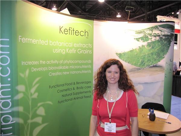 A woman with long curly hair in a red top and lanyard stands in front of a green Kefitech banner about fermented botanical extracts using kefir grains, near a small round table.