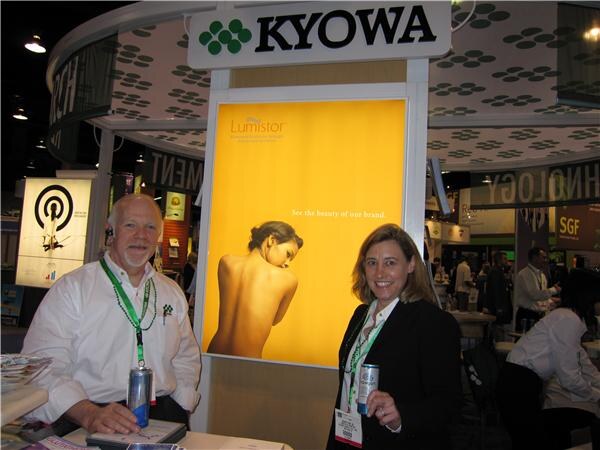 A bearded man in a white shirt and a woman in a dark blazer, both with lanyards and holding beverage cans, stand beside a yellow Lumistor lightbox under a Kyowa sign.