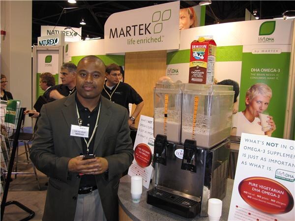 A man in a dark blazer with a lanyard and name badge stands holding a phone in front of the Martek life's DHA booth, beside a Horizon Organic milk carton and a beverage dispenser.